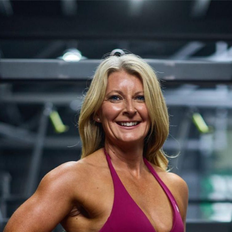 Fit woman in a gym wearing a pink sports outfit with weights and exercise equipment in the background.