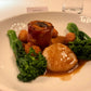 A plated gourmet dish on a white plate, featuring a glazed meat cylinder, a seared scallop, broccoli, and small fried balls, set on a restaurant table with blurred diners in the background.