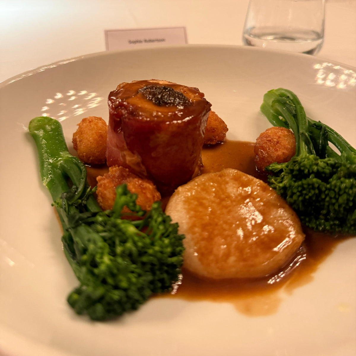 A plated gourmet dish on a white plate, featuring a glazed meat cylinder, a seared scallop, broccoli, and small fried balls, set on a restaurant table with blurred diners in the background.