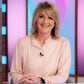 A smiling blonde  Kaye adam  - woman wearing a light pink long-sleeve blouse sits behind a clear desk in a brightly lit studio with pink and blue background panels.