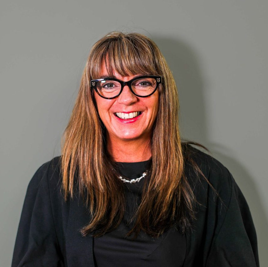 A smiling woman with long brown hair and black-rimmed glasses, wearing a black top and a silver necklace, photographed against a plain gray background.