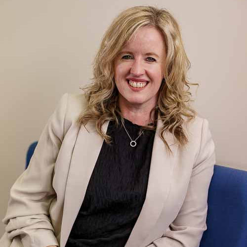 Woman in a beige blazer sitting on a blue chair against a plain background