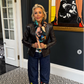 Woman standing in a room with a black wall and patterned rug, wearing a black leather jacket and colorful scarf.